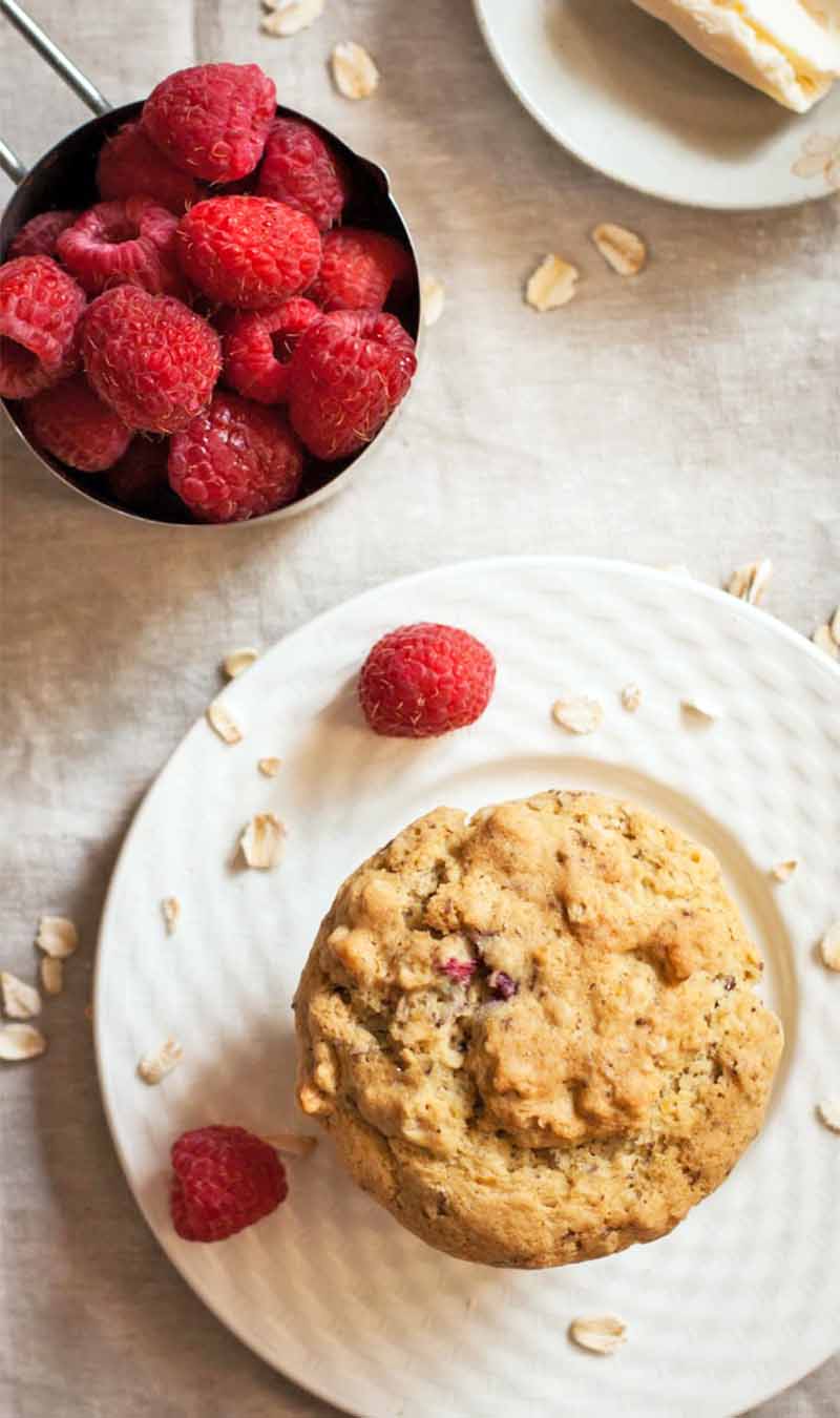 Vertical top-down image of a light brown muffin on a plate beside a small dish of butter and a silver measuring cup of berries, with scattered oats and berries on a beige cloth background.