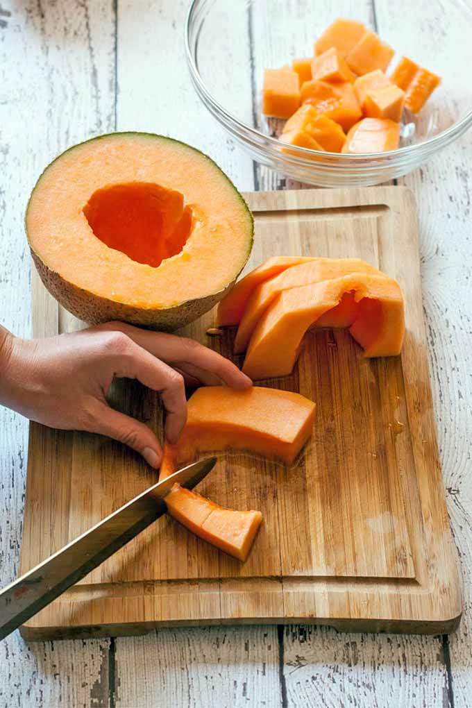 Half of a cantaloupe rests on a wooden cutting board next to a woman's left hand that is holding a slice of the orange flesh and cutting it with a chef's knife held in her other hand, next to several slices lined up in a row, and a glass bowl of chunks of the fruit, on a white rustic wooden table.
