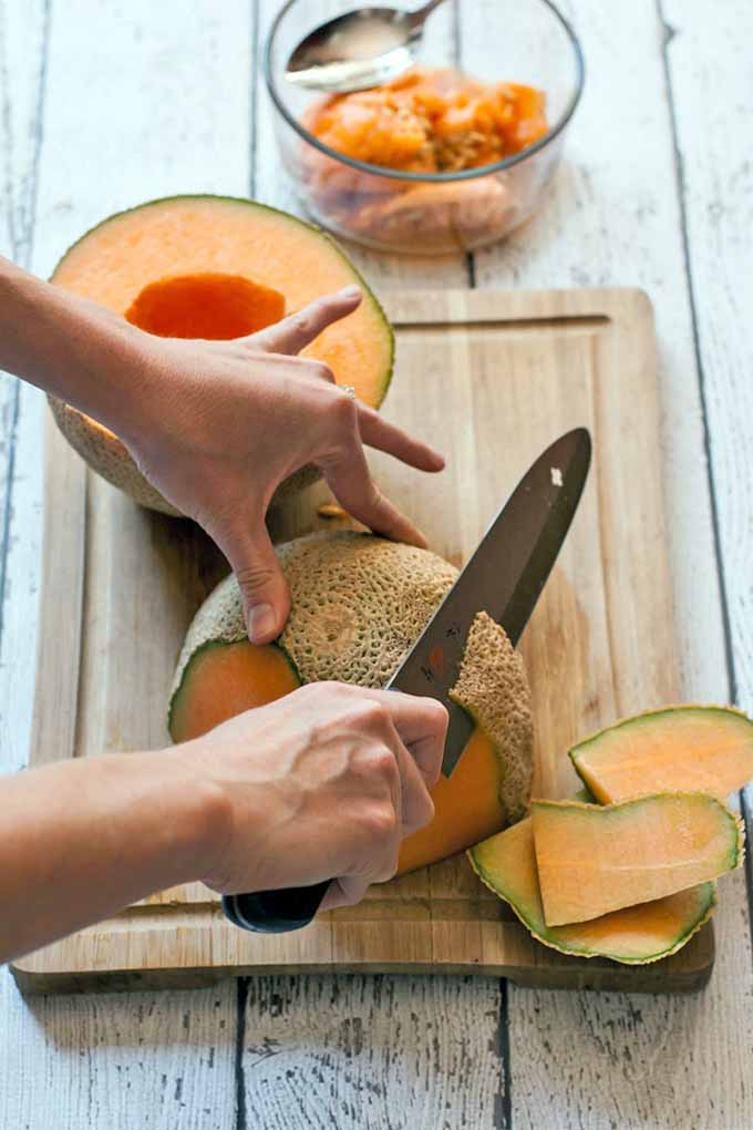 Closeup of a woman's hands peeling half of a cantaloupe, with the other half sitting beside it on a wooden cutting board, with a glass bowl towards the top of the frame that contains the seeds of the melon.