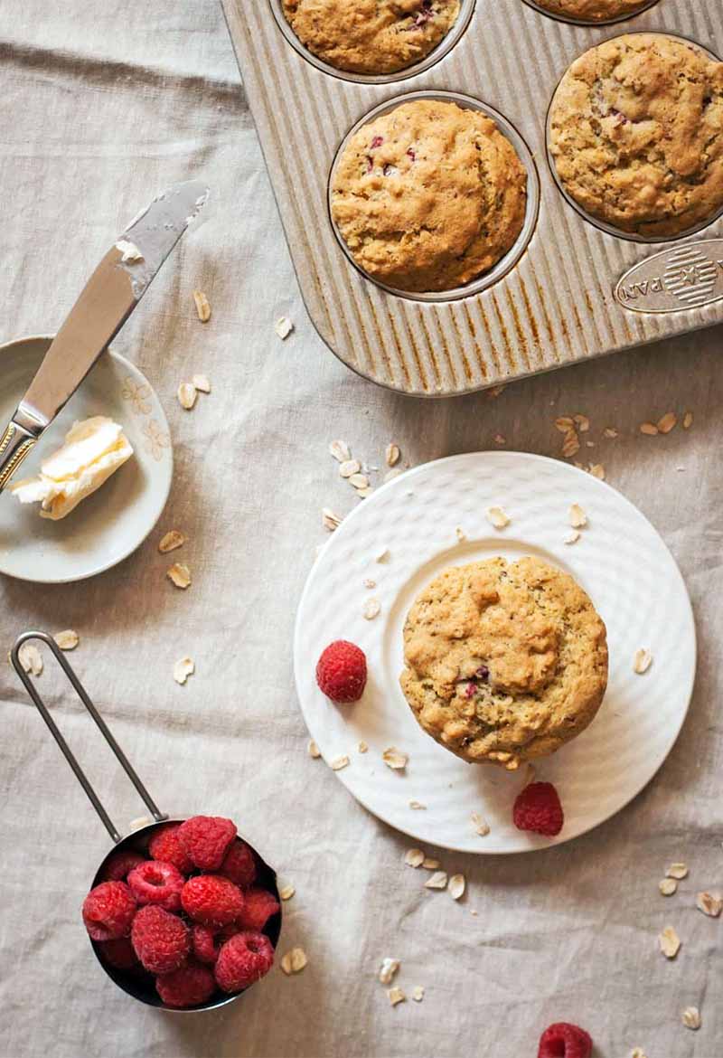 Vertical top-down image of a tin of freshly baked muffins beside another on a small white plate with two raspberries, a dish of butter with a knife, and a stainless steel measuring cup of berries, on a beige cloth with scattered oats.
