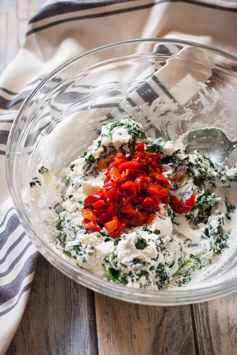 Ricotta, kale, chopped red peppers and rosemary being blended in a glass mixing bowl
