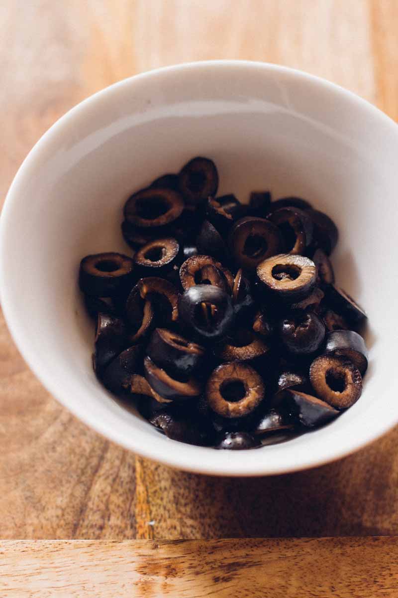 A close up of a bowl of sliced black olives sitting on a medium wood table.