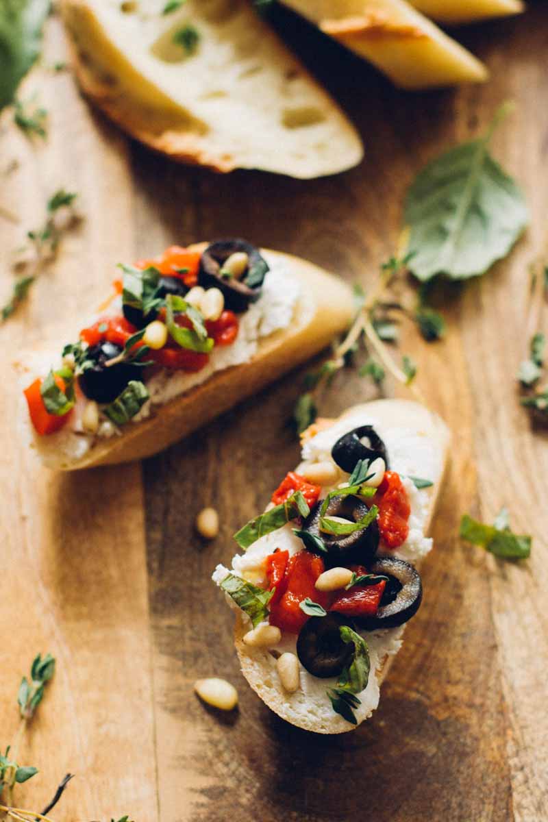 Top down view of a two slices of baguette bread made into crostini topped with red peppers, black olives, goat cheese, pine nuts, and basil olive oil.