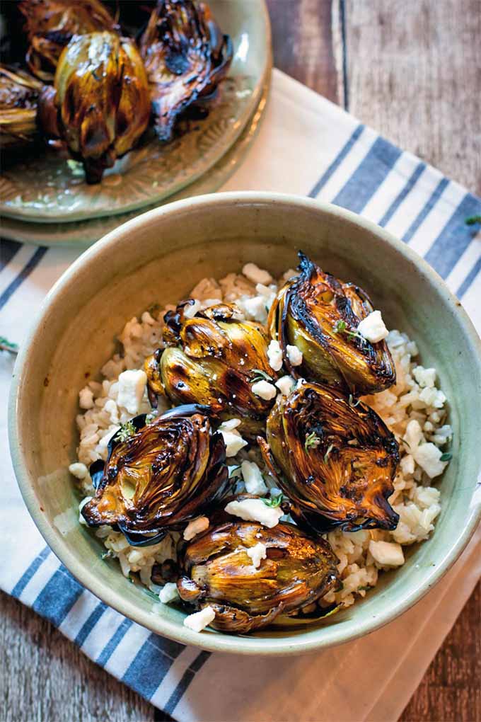 Top-down shot of a plate of grilled baby artichokes and more of the same topping a bowlful of brown rice and crumbled feta, on a blue and white striped cloth atop a brown wood table.