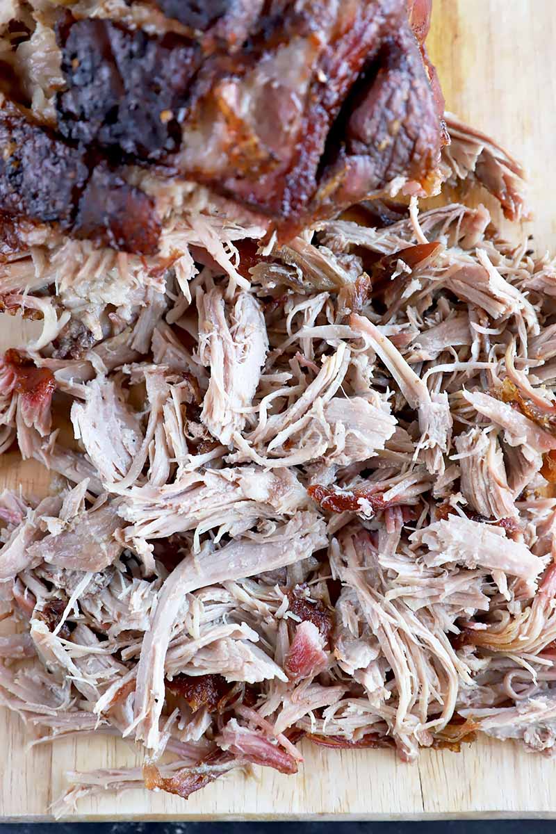 Vertical top-down image of shredded a large chunk of cooked meat on a wooden board.