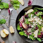Top-down shot of a black bowl of salad with sliced apple, half a head of radicchio, a fork and serrated knife, and scattered greens on a gray tablecloth.