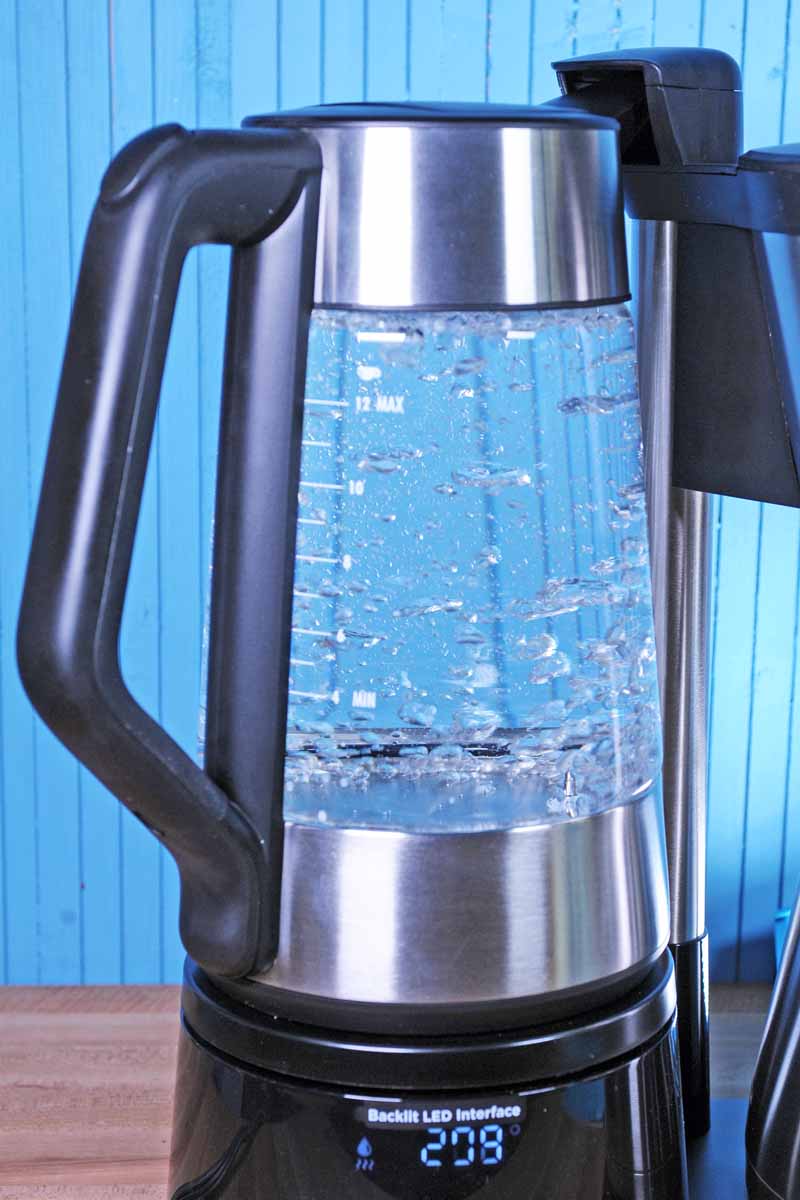 A close up of the borosilicate glass kettle on the OXO On 12 Cup Coffee Maker. Water is starting to boil inside of the kettle. Background is a medium blue painted wood with back lighting.