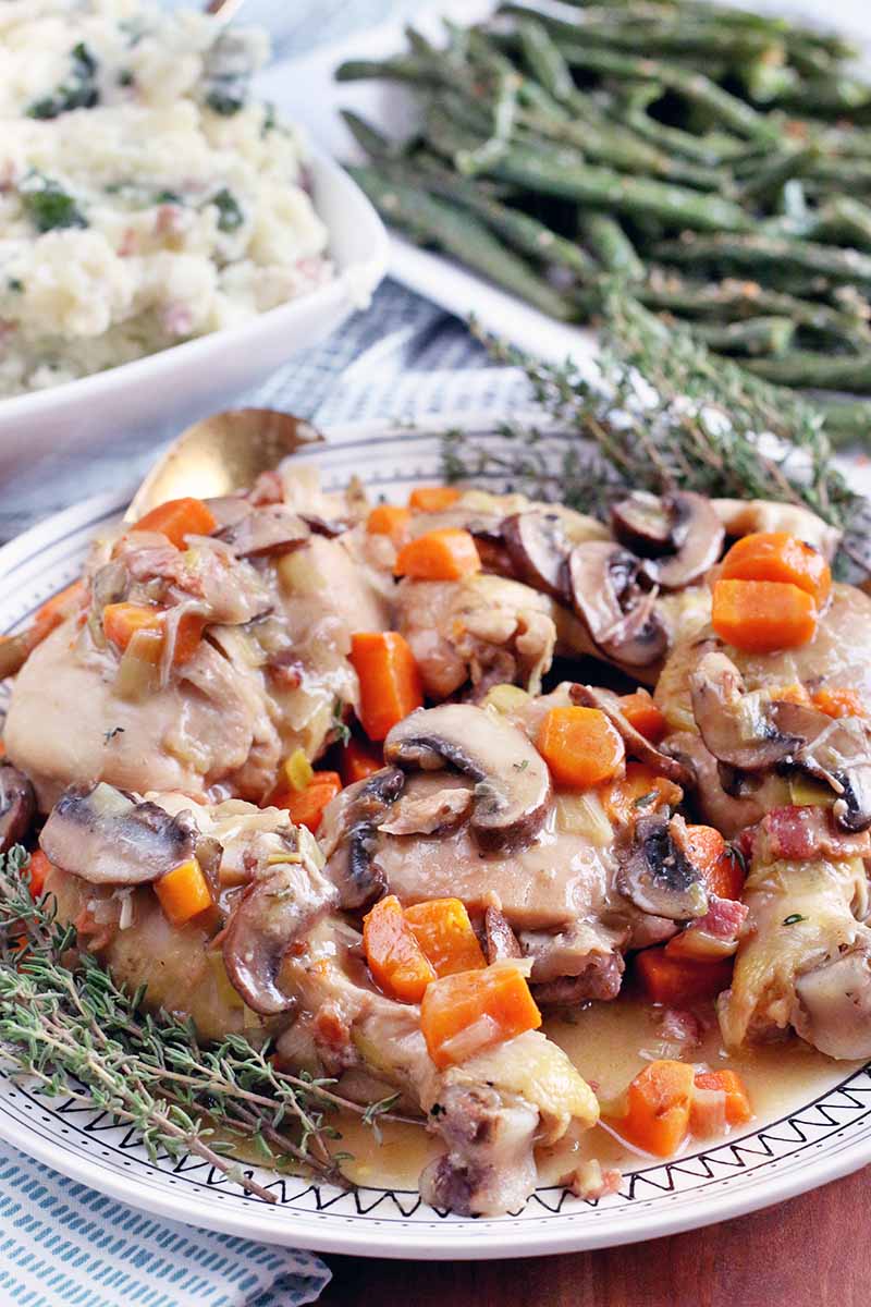 Vertical image of a serving platter of braised chicken with carrots and mushrooms in the foreground, garnished with sprigs of fresh thyme, with a bowl of kale mashed potatoes and a platter of parmesan green beans in the background.