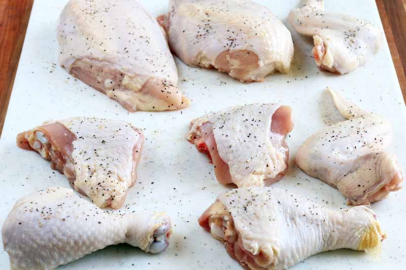 Raw chicken pieces arranged on a white cutting board, sprinkled with salt and pepper.