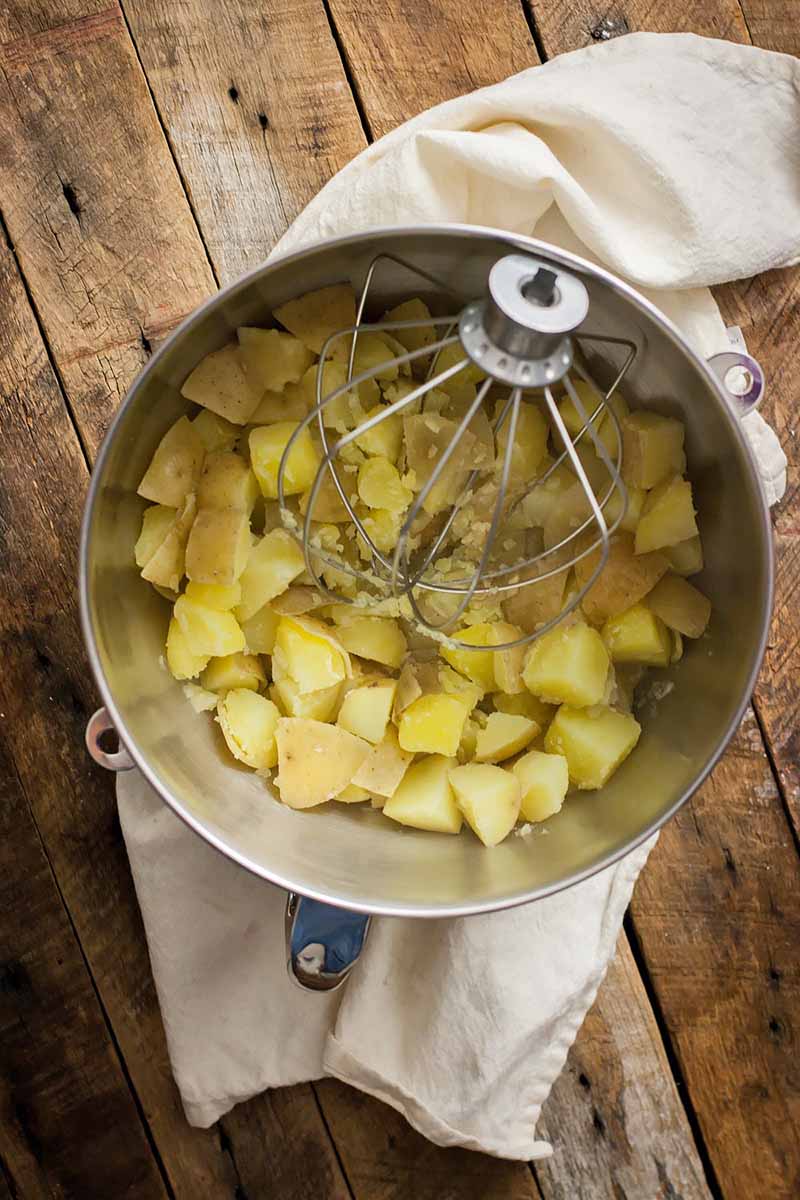 Vertical image of a metal bowl with chunks of potatoes and a wire whisk.