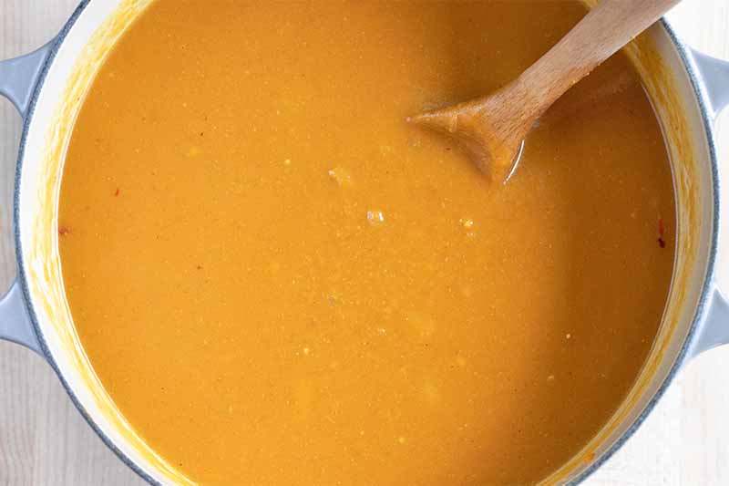 Overhead shot of a light blue enameled pot filled with an orange blended soup, with a wooden spoon stuck into it, on a beige background.