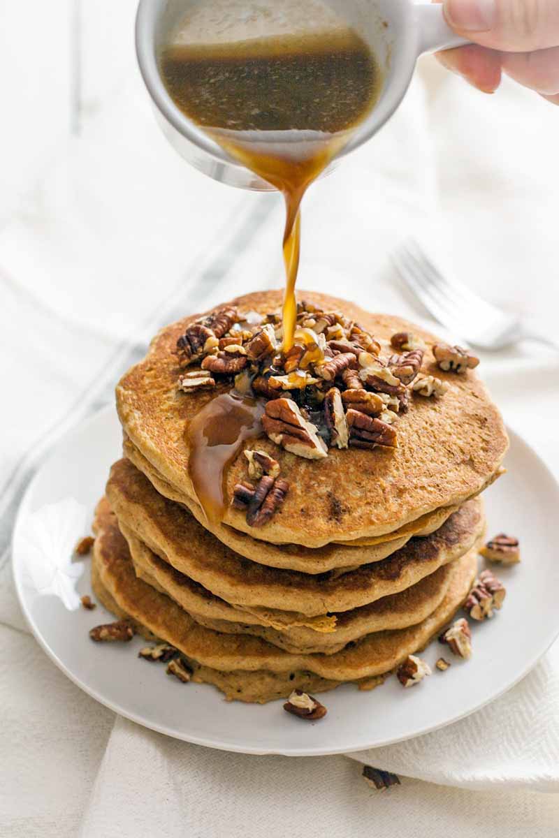 A hand drizzles a brown syrup from a metal measuring cup onto a stack of pancakes below that are topped with chopped nuts, on a white plate, with a white cloth background.