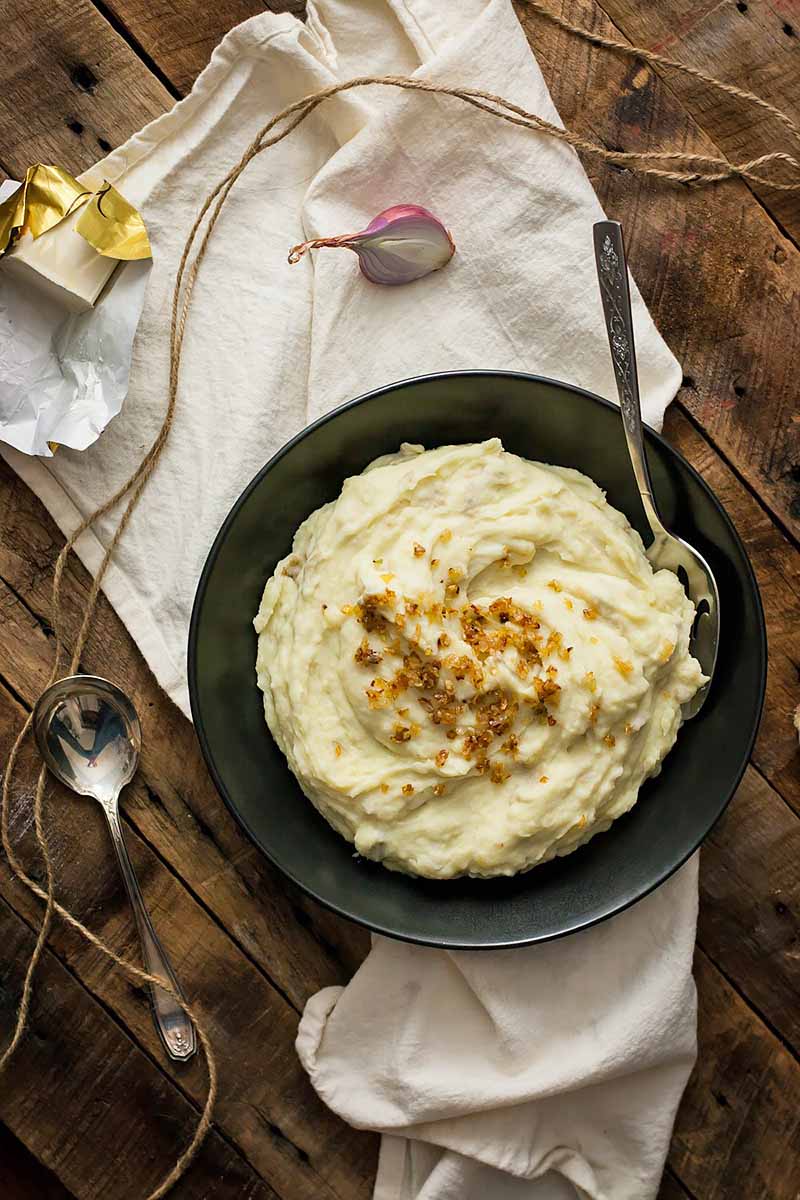 Vertical image of a bowl of mashed potatoes in a black bowl on a white towel with metal spoons and shallots and kitchen twine.