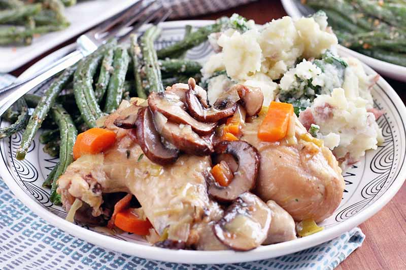 A dinner plate is piled high with wine braised chicken with vegetables, parmesan green beans, and kale red-skinned mashed potatoes, with a fork.