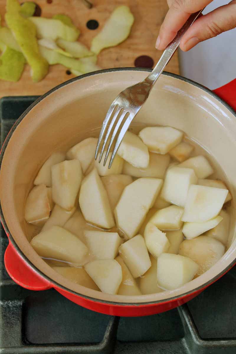Peeled and sliced pears are poaching in the bottom of a red enameled pot on a black stove, and a hand holds a fork towards the contents of the pan, about to check for doneness, with a wooden cutting board with green fruit peels on top in the background.