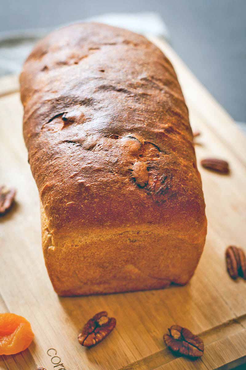 A golden brown loaf of homemade bread on a wood cutting board, with scattered pecans and dried apricots, on a gray cloth background.