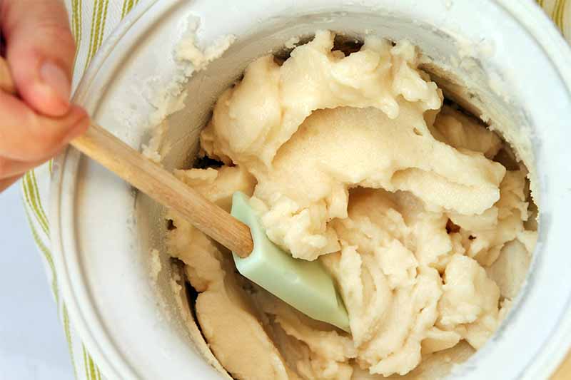 Closely cropped overhead shot of a hand stirring a beige-colored sorbet in an ice cream maker freezer canister with a mint-colored rubber spatula with a wood handle.