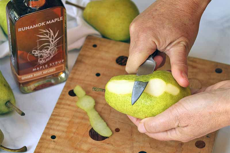 Closeup of hands holding a pear in the left and peeling it with a paring knife with the right, with a wooden cutting board, whole fruit, and a bottle of maple syrup in the background.