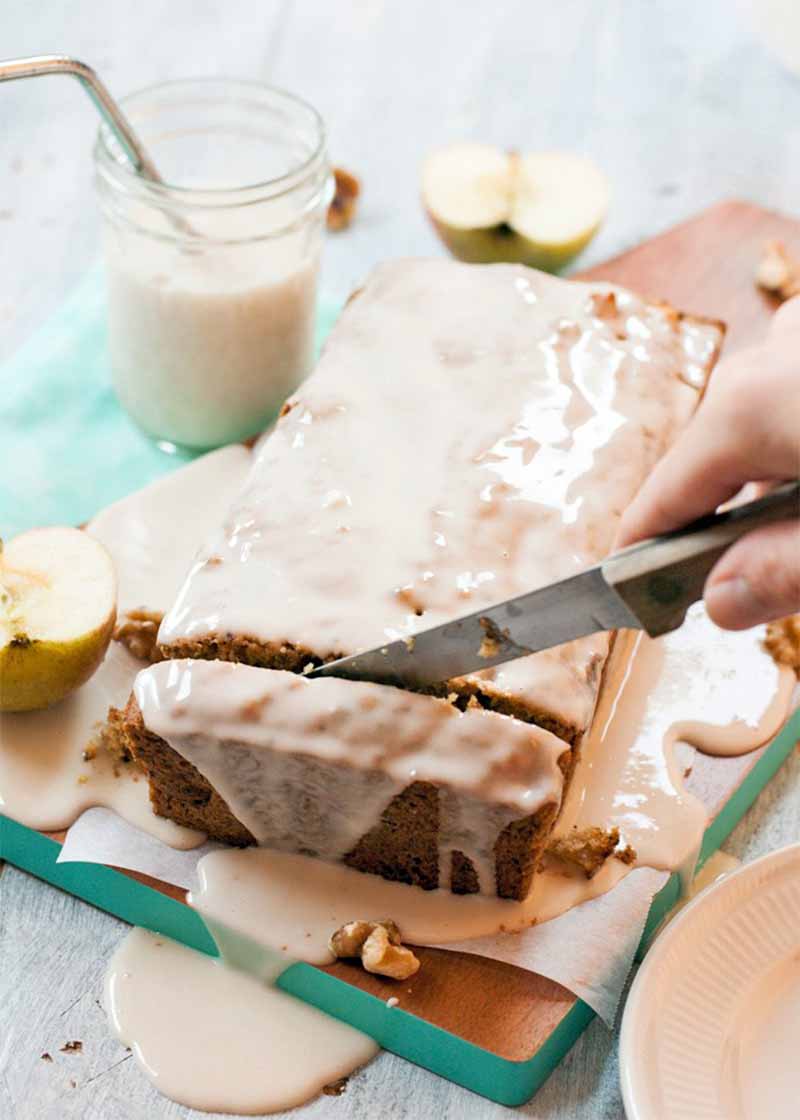 A hand holds a knife and slices into a brown loaf-shaped vegan cake topped with a thin, white sugar icing, with a cut apple and a mason jar of almond milk on a light aqua cloth surface.