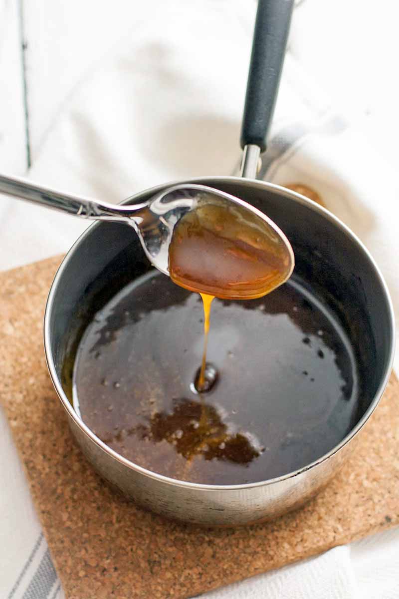 A spoon drizzles a brown liquid syrup into a small saucepan on a brown cork trivet, on top of a folded white kitchen towel.