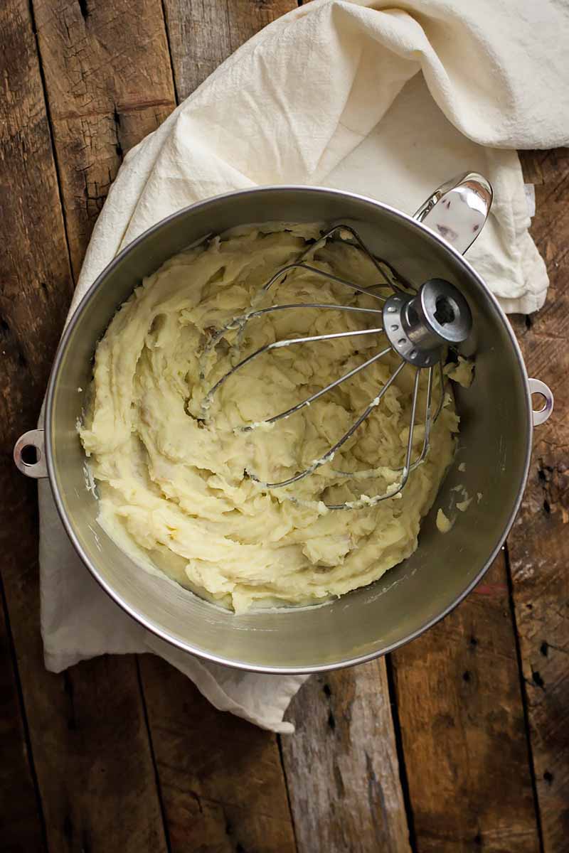 Vertical image of mixing mashed spuds in a metal mixing bowl with a wire whisk.