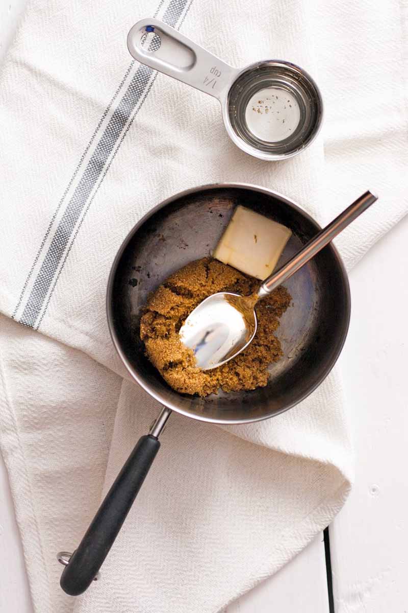 Overhead shot of a small metal measuring cup and a small nonstick saucepan with margarine and brown sugar at the bottom and a spoon resting on top, on a folded white kitchen towel with a blue stripe.