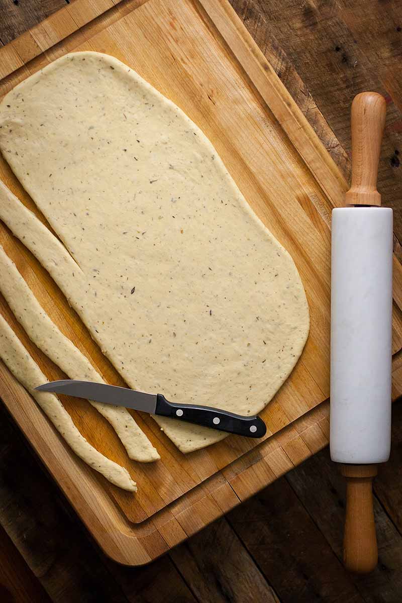 Top-down view of a bread dough rolled out onto a cutting board and partially cut into strips. A kitchen knife is to the lower left and a marble rolling pin sits to the right of the frame.