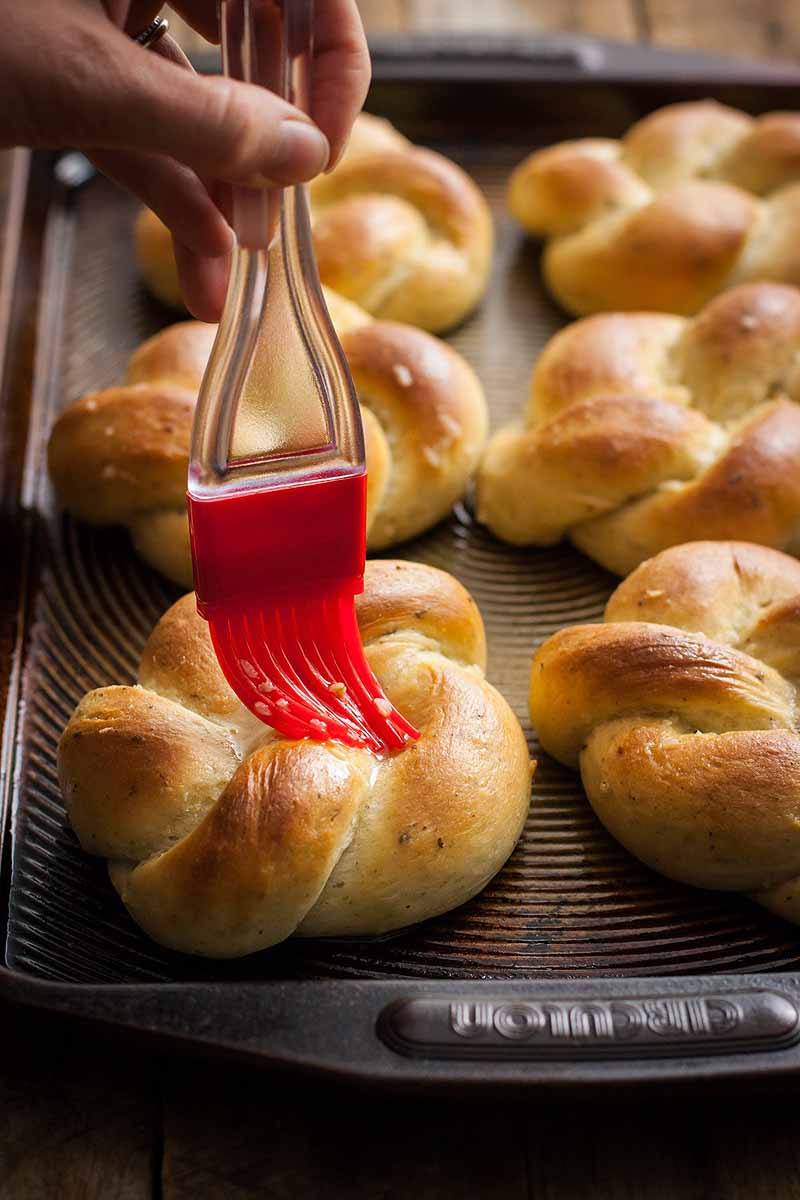 A silicone brush being used to spread vegan margarine over the top of a roasted garlic knot dinner roll.
