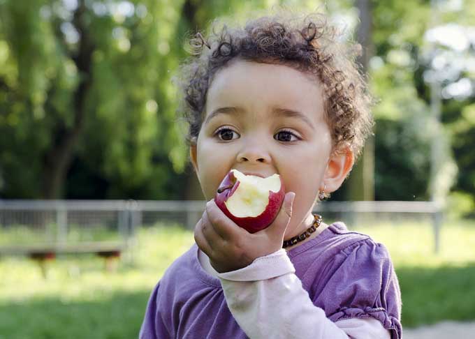 A small child eating an apple outdoors, with trees and grass in soft focus in the background.
