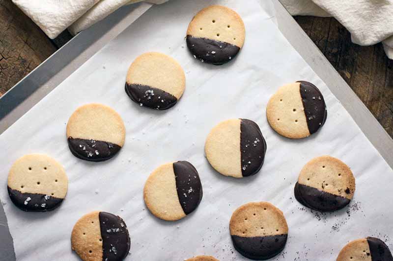 A rimless baking sheet lined with parchment paper and topped with chocolate-dipped shortbread cookies, arranged at a diagonal, on a brown wood surface with a folded off-white cloth.