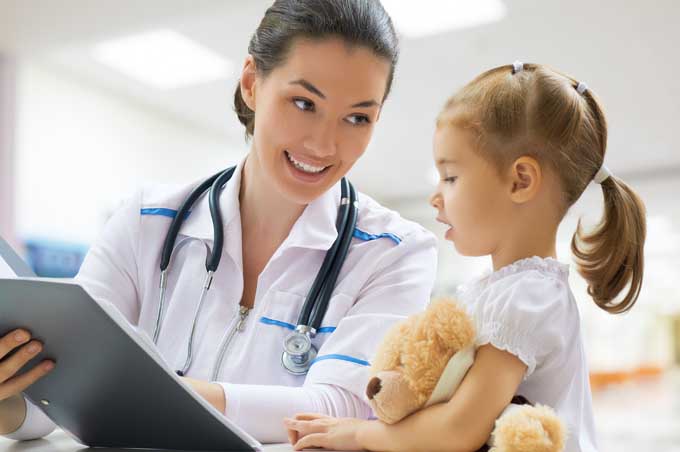 A doctor wearing a stethoscope and holdig a clipboard talks to a young girl with a ponytail holding a teddy bear.