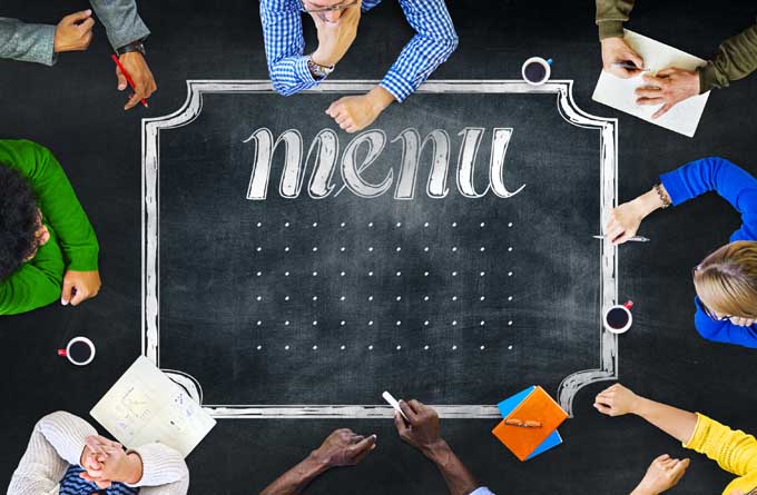 Overhead closely cropped image of people sitting around a large chalkboard, with just their arms visible and two people's heads at the right and left sides, with a chalkboard menu and a grid of dots printed on the board.