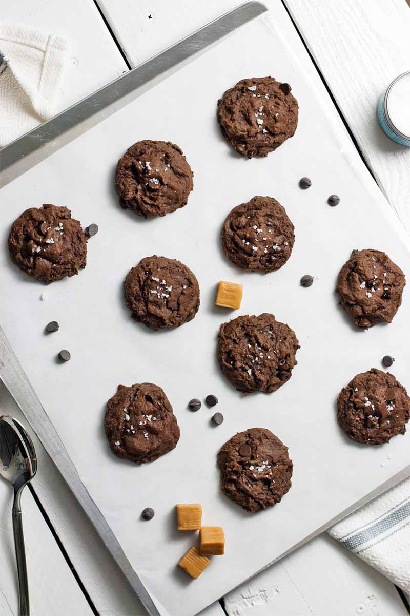Top-down shot of 10 chocolate cookies topped with sea salt, on a parchment-lined baking sheet with scattered caramel candies and chocolate chips, on a white wood background with a spoon to the left and a canister at the top right.