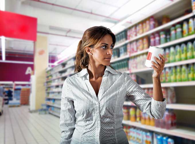 A woman examines a food label on a can in an aisle of the grocery store.