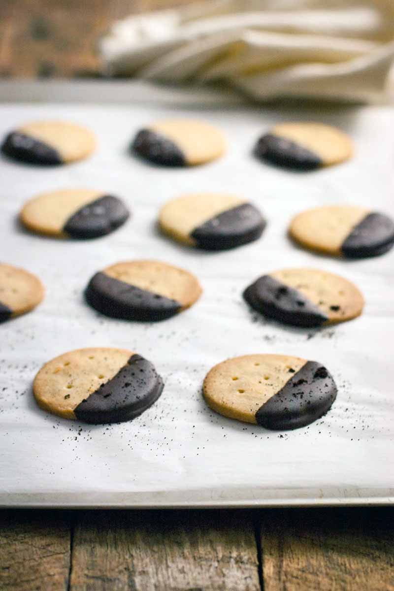 Closely cropped vertical shot of round cookies with holes poked in the top, arranged on a parchment-lined baking sheet after they have been dipped in chocolate and sprinkled with espresso powder.