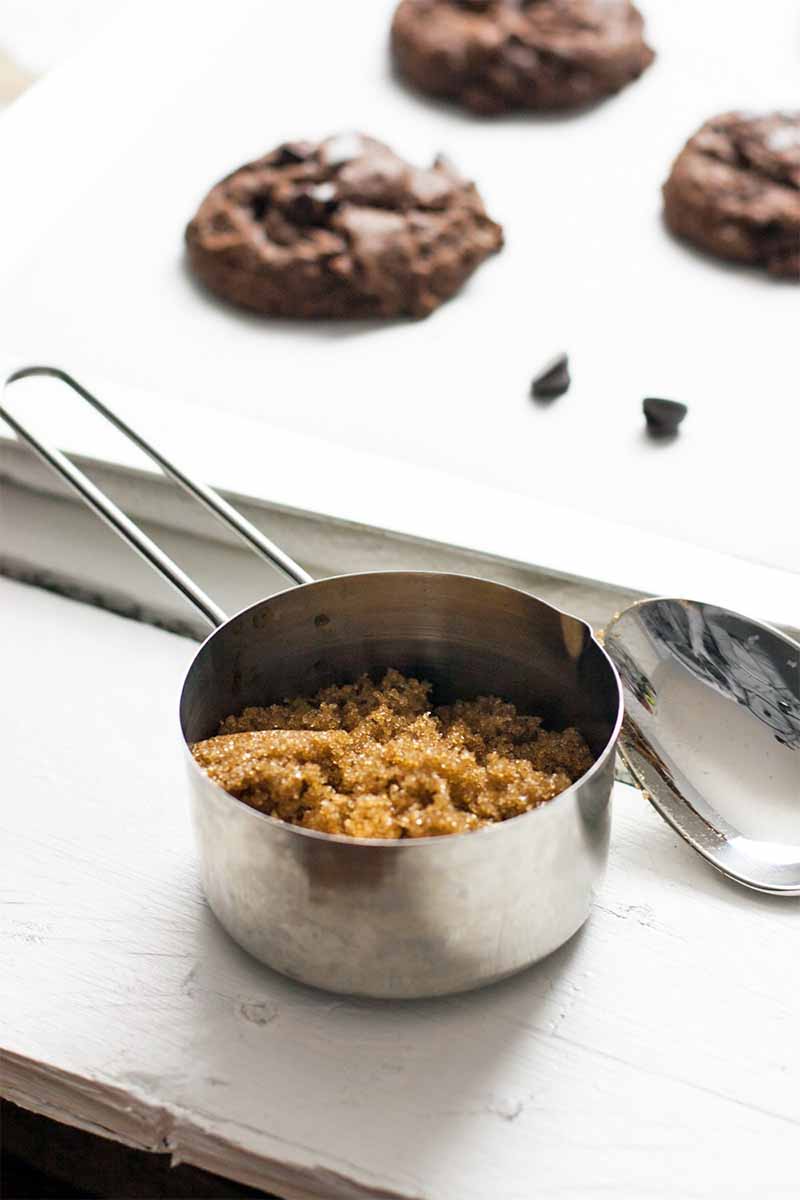 A stainless steel measuring cup of brown sugar with a spoon in the foreground, and a parchment-lined sheet pan topped with homemade cookies and scattered chocolate chips in the background.