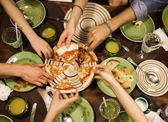 Overhead shot of six hands reaching for slices of pizza on a plate, surrounded by more cups and dishes on a dinner table.