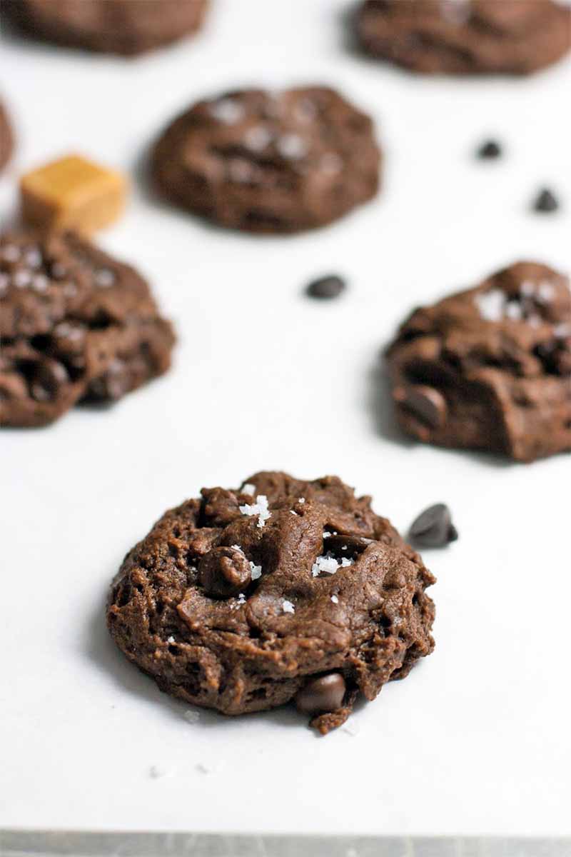 Three rows of chocolate cookies sprinkled with sea salt, with a caramel candy and a few chocolate chips on a white background, shot at an oblique angle.