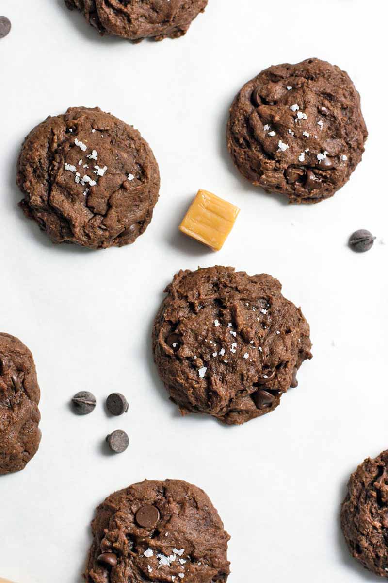 Top-down shot of chocolate cookies sprinkled with salt, with a square caramel candy and scattered chocolate chips, on a white background.