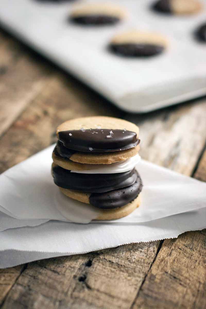 Vertical image of a short stack of round vegan short cooked, some of which are dipped in chocolate and others coated in a white glaze, on a folded piece of parchment paper on top of a brown wood surface, with more cookies on a parchment-lined baking sheet in soft focus in the background.