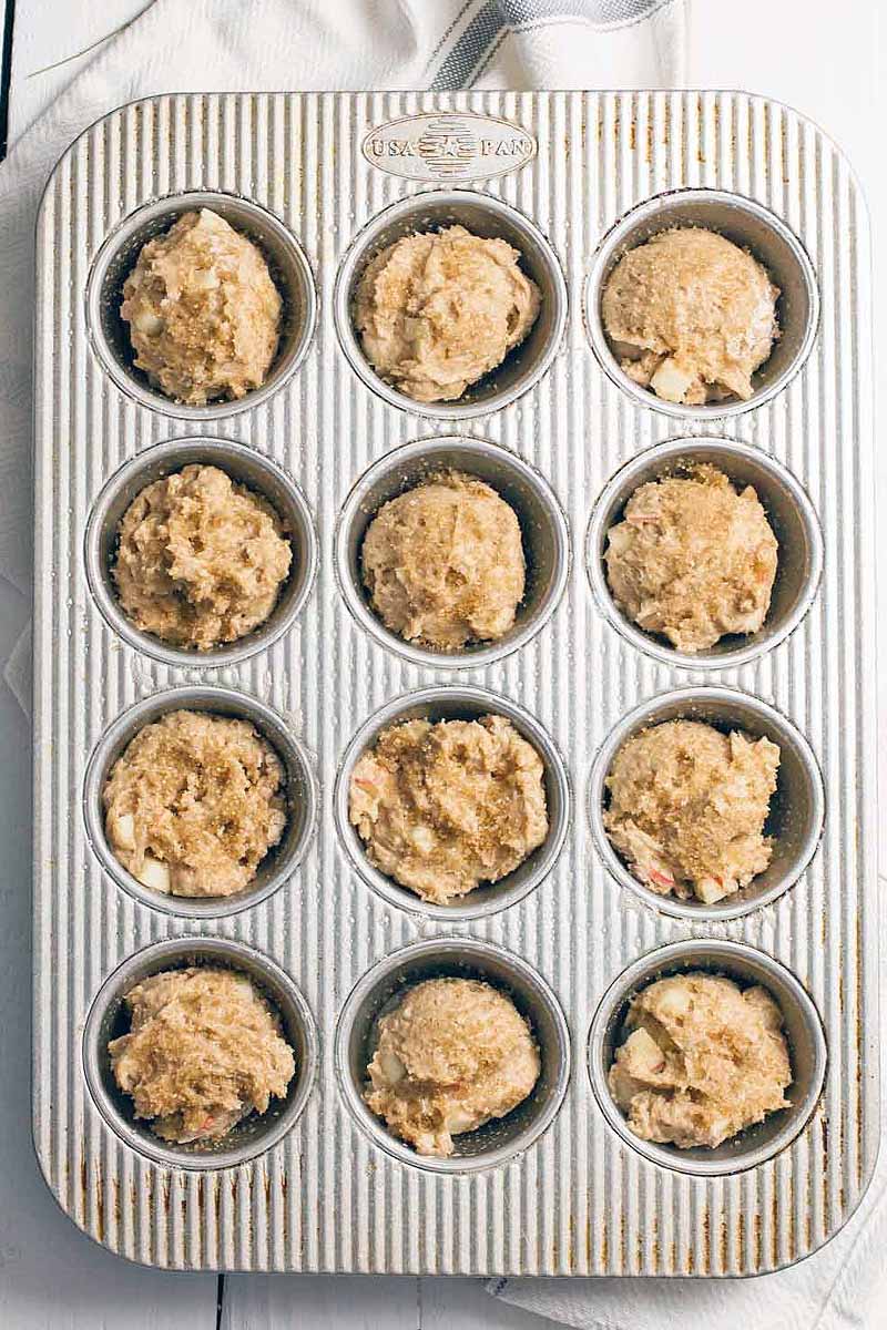 Top-down shot of a muffin tin with twelve holes that are filled with a tick brown batter, on a white background with a white dish towel with a blue stripe.