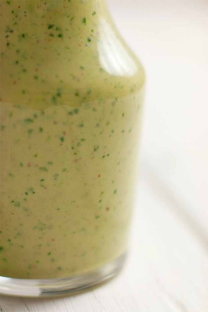 Vertical image of a glass salad dressing bottle of green cilantro and cayenne tahini sauce, on a white background.