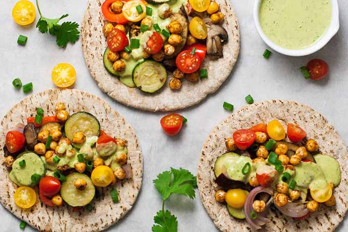 Three pieces of pita bread with roasted and fresh vegetables and garbanzo beans on top, with a small white ceramic dish of a pale green sauce, and scattered red and yellow cherry tomatoes and green cilantro leaves, on a white piece of parchment paper.