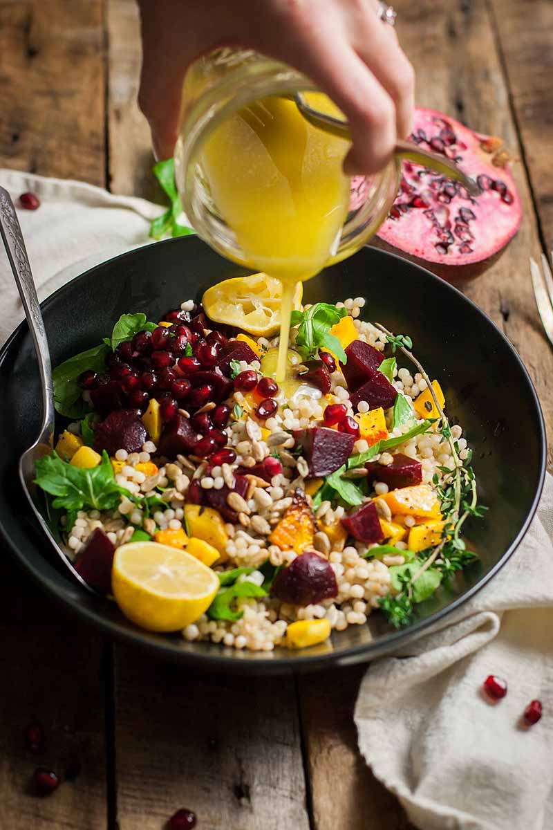 A human hand pours a lemon dressing over a Roasted Butternut Squash and Beet Couscous Salad.