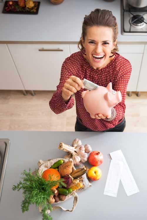 A woman puts a folded bill into a piggy bank, smiling at the camera, standing in a galley kitchen with fresh vegetables, fruit, and herbs on a gray countertop.