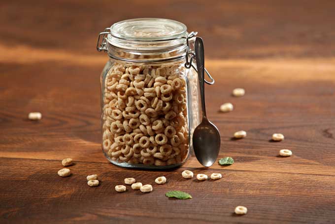 Closeup image of a swing-top glass jar of Cheerios, with a metal spoon, on a wood surface with scattered pieces of cereal and two mint leaves.
