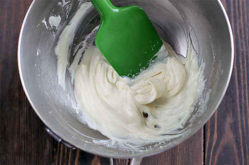 Melted white chocolate in a stainless steel bowl, being stirred with a green silicone spatula, on a dark brown wood background.