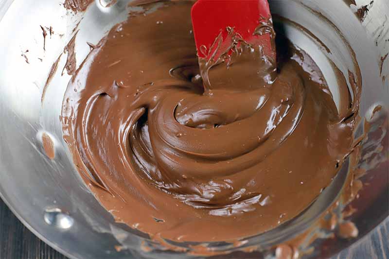 Melted milk chocolate being stirred with a red silicone spatula, in a stainless steel mixing bowl.