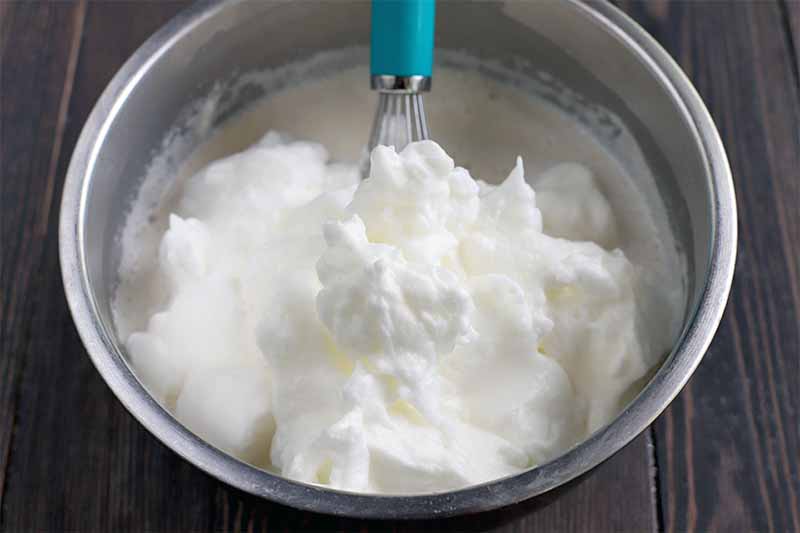 Egg whites whipped to form still peaks in a stainless steel mixing bowl, with a blue and silver whisk, on a dark brown wood surface.