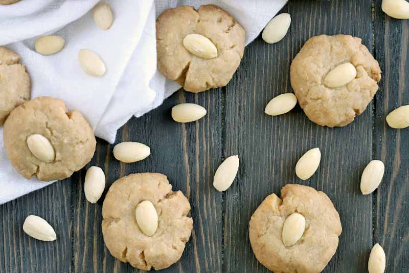 Brown butter cookies and blanched almonds scattered on a brown wood surface that's partially covered with a white cloth.